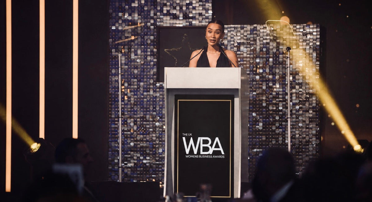 A woman stands behind a white podium with a large black front panel reading WBA, at what appears to be a formal event or awards ceremony. The stage has a dark mirrored tile backdrop, vertical light bars, and yellow spotlights, with an audience seated in the foreground.